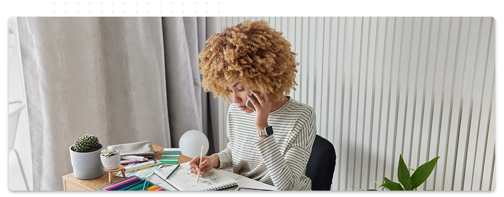 Une jeune femme, concentrée, dessine dans un carnet tout en parlant au téléphone, assise à un bureau avec des fournitures de dessin colorées, dans un espace de travail moderne et lumineux.