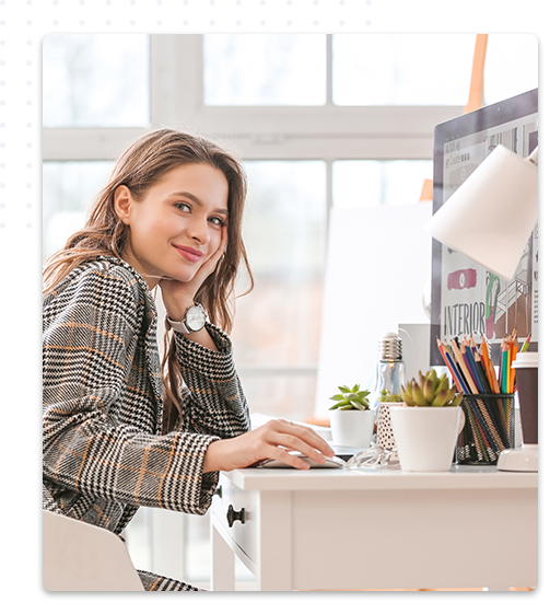 Une jeune femme souriante, vêtue d'une veste à carreaux, travaille à son bureau sur un ordinateur, avec un écran affichant un projet de décoration intérieure. Le bureau est décoré de plantes succulentes et de fournitures colorées, dans un espace lumineux avec une grande fenêtre en arrière-plan