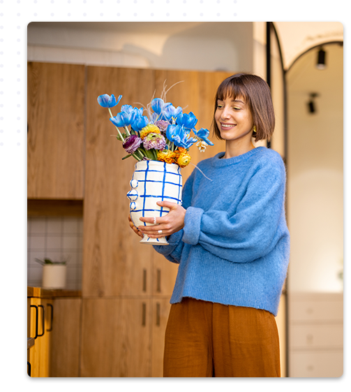 Une décoratrice d'intérieur souriante, habillée d'un pull bleu, tient un vase rempli de fleurs colorées dans une cuisine moderne avec des éléments en bois naturel.