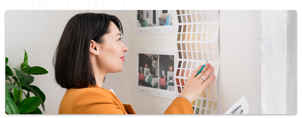 Une décoratrice d'intérieur souriante consulte des échantillons de couleurs et de tissus étalés sur une table, dans un espace de travail moderne et lumineux avec des plans et des croquis accrochés au mur en arrière-plan.