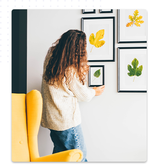 Une décoratrice d'intérieur ajuste un cadre photo représentant des feuilles encadrées sur un mur, créant une galerie murale dans un espace de vie. Un fauteuil jaune est visible au premier plan.