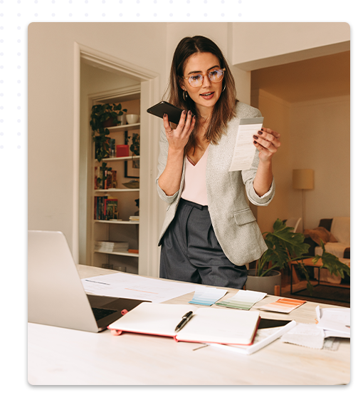 Une décoratrice d'intérieur, portant un blazer gris, parle au téléphone tout en tenant des documents, debout à côté d'un bureau où sont étalés des échantillons de couleurs et des papiers, symbolisant une activité de home staging.