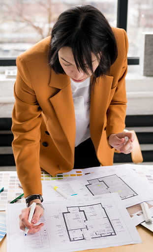 Une femme, décoratrice d'intérieur, vêtue d'un manteau orange travaille sur des plans architecturaux étendus sur une table, dans un bureau lumineux avec une vue sur la ville en arrière-plan.